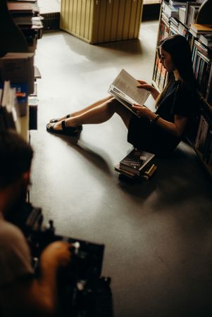 Side view of young woman sitting between bookshelves reading a book