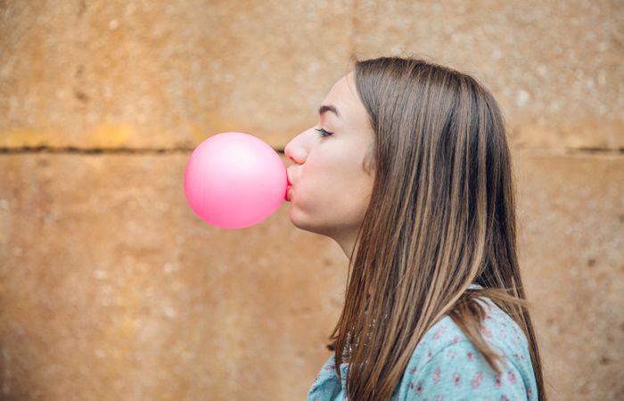 Side view of female standing outside in front of stone wall blowing bubble with pink gum