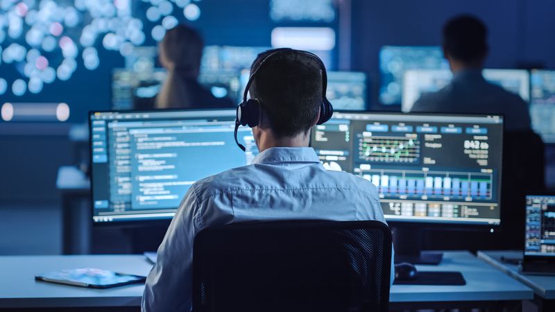 IT Specialist With Headset Monitors Real-time Data on Multiple Screens in Blue-lit Operations Center. Colleagues Work in Background Amid Displays Showing Complex Information.