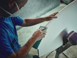 Carpentry man in respirator polishing surfboard with abrasive