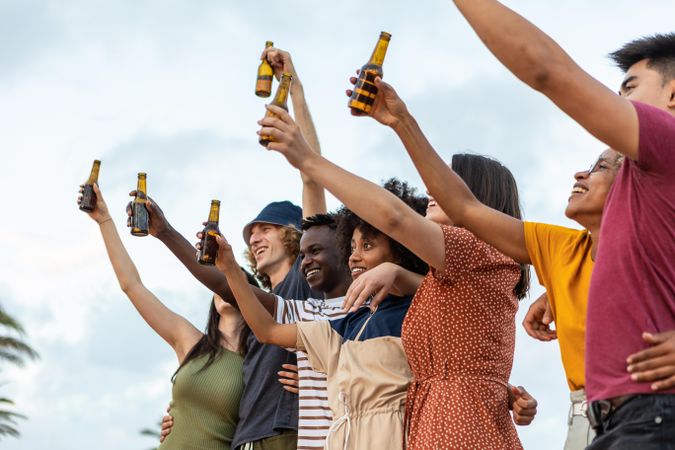 Group of multiracial friends raising arms while drinking beer at summer party