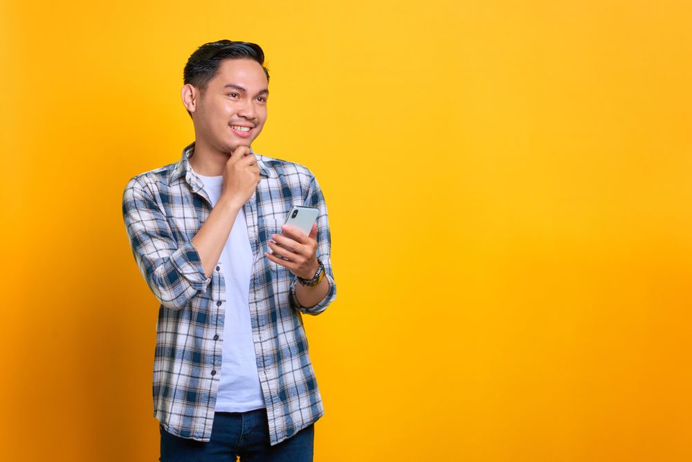 Smiling Asian male holding his phone with hand on his chin