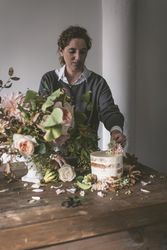 Woman placing plate with cake decorated flower on table with bouquet of blooms in vase