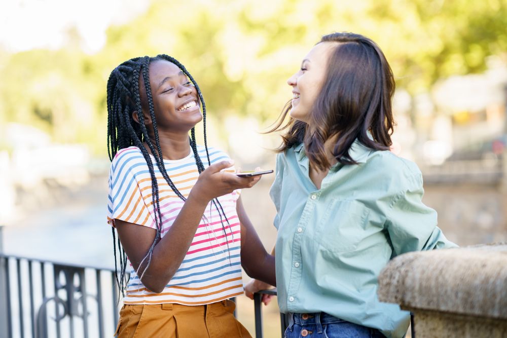 Female friends laughing while using speaker phone in casual clothing outside