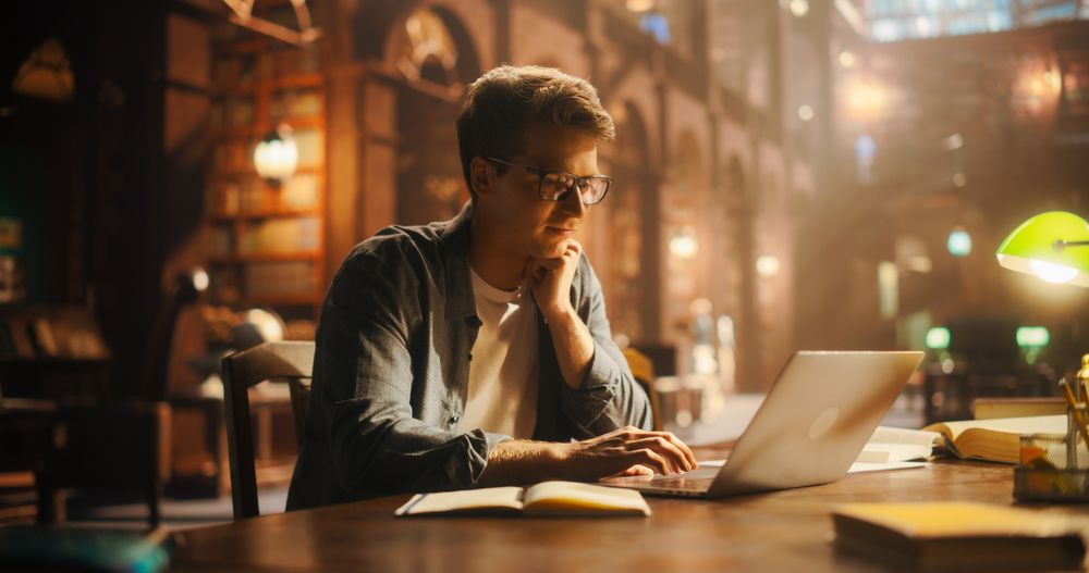 Concentrated Young Male Student Engaged in E-Learning With Laptop in a Vintage Library Setting