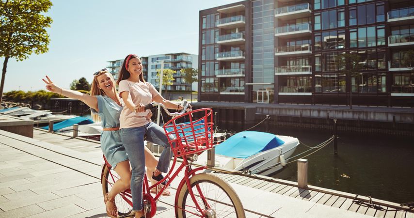 Two happy female friends laughing while riding bike along the riverwalk