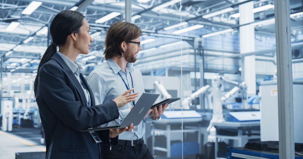 Portrait of Cheerful Engineer and Happy Asian Scientist Using Laptop and Tablet Computers, Talking in a Factory Facility with Robots Producing Modern Electronic Components for Different Industries