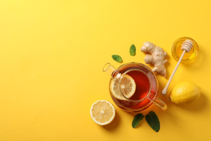 Flat lay. Teapot, lemon, ginger, mint, honey and dipper on yellow background, copy space