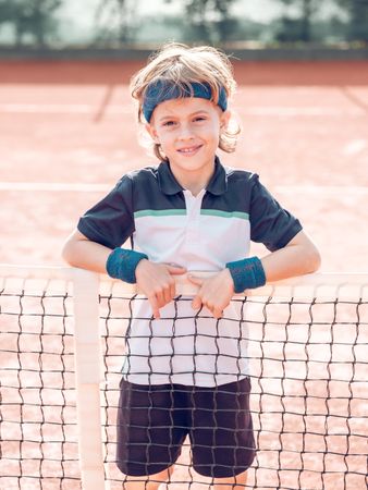 Cute sporty kid smiling at camera while standing on tennis court