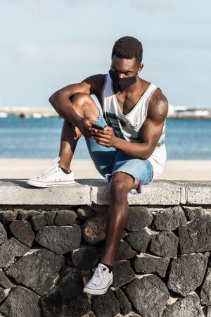 Man in tank top and blue shorts sitting on concrete wall working on his phone