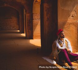 Man in traditional outfit sitting on floor leaning on pillar 423P7b