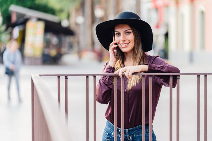 Charming woman in trendy outfit talking on smartphone and smiling at city street