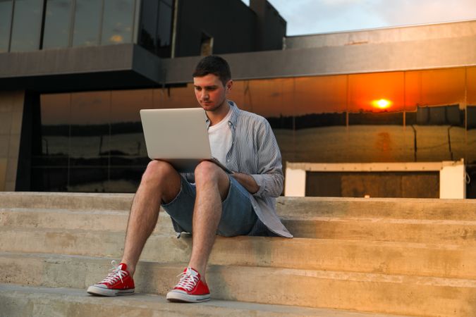 Young man sits in steps and working in laptop outdoor. Freelancer