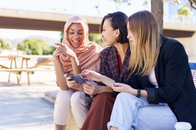 Three happy women sitting on outdoor park bench talking while watching smartphone