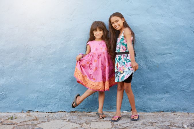 Full length portrait of two happy girls standing by a blue wall