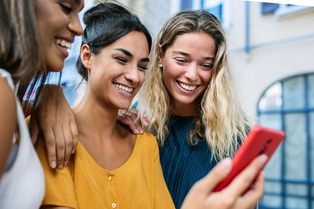 Three young diverse women smiling while using mobile phone together in city street