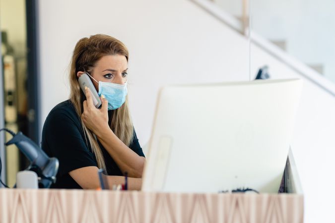 Woman in dark shirt wearing facemask using computer and talking on the phone