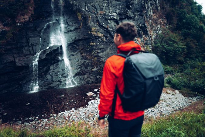 Back view of a man in red jacket with backpack standing near waterfalls