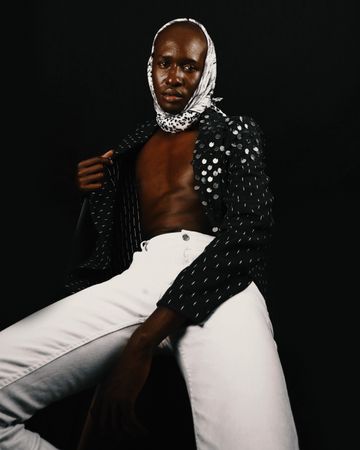 Bare-chested man in polka dot top and headscarf sitting against dark background