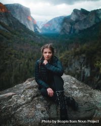 Young woman in dark bubble jacket sitting on rock formation near lake 0P98g5