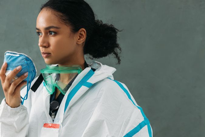 Side view of Black female medical worker with PPE goggles around her neck holding a face mask