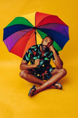 Happy Black male thinking about something while sitting under colorful umbrella
