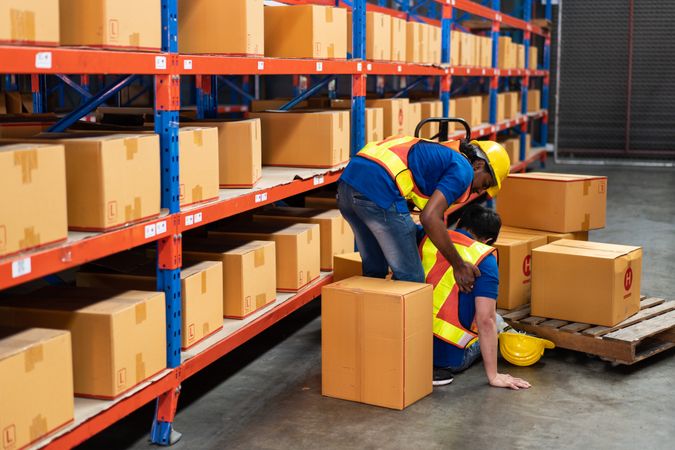 Workplace accident in a warehouse, a worker helping an injured colleague on the floor.