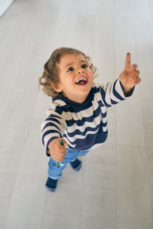 Cheerful kid in striped hoodie pointing up at home