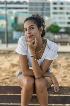 Young woman in light shirt and denim short sitting on wooden bench outdoor