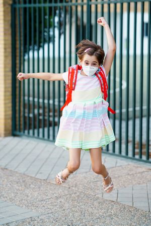 Excited girl jumping outside school gates wearing facemask