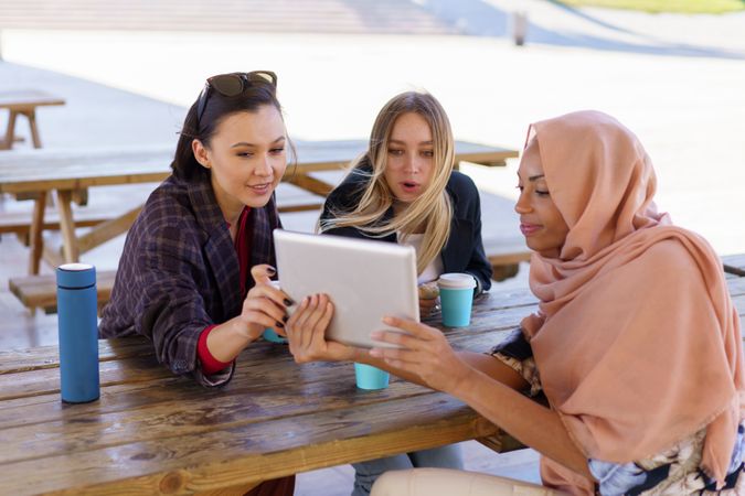 Three women on park bench with insulated mugs and digital tablet
