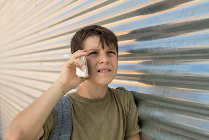 Serious boy talking on smartphone leaning on metal shutter