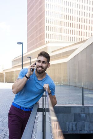 Smiling man leaning on glass railing outside having conversation on mobile phone