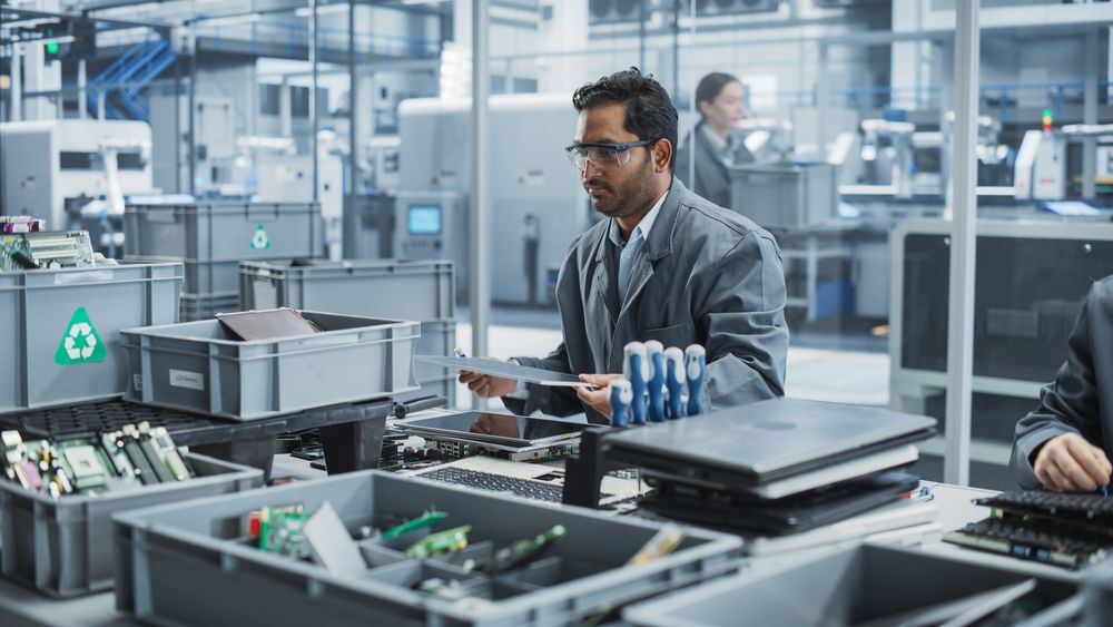 Indian Male Worker Disassembling Old Laptops To Recycle Electronic Components For Microchip Production At an Automated Electronics Factory With Robotic Arms.