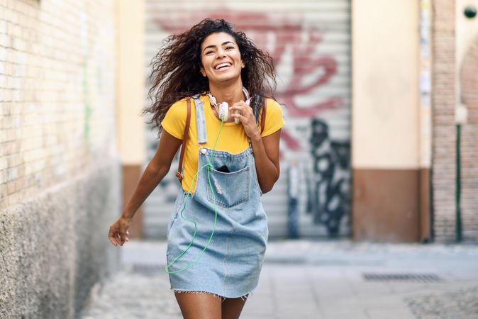Joyful Arab woman with headphones and curly hairstyle walking outdoors in lane with graffiti