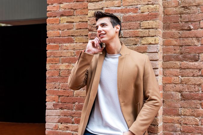 Male in camel coat talking on phone leaning on brick wall