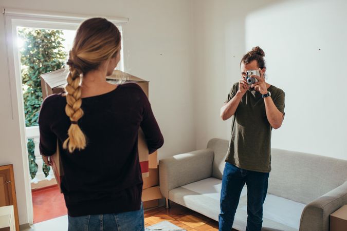 Man taking a photograph of a woman carrying a packing box in new home