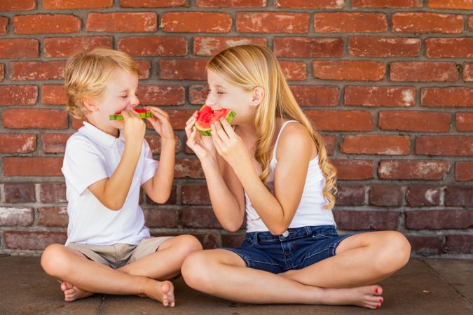 Cute Young Boy and Girl Eating Watermelon Against Brick Wall