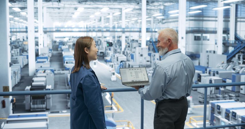 Mature Engineer and Young Japanese Technician Having a Conversation and Using Laptop Computer. Specialists Monitoring, Analyzing Conditions at a Modern Electronics Factory with Automated Robots
