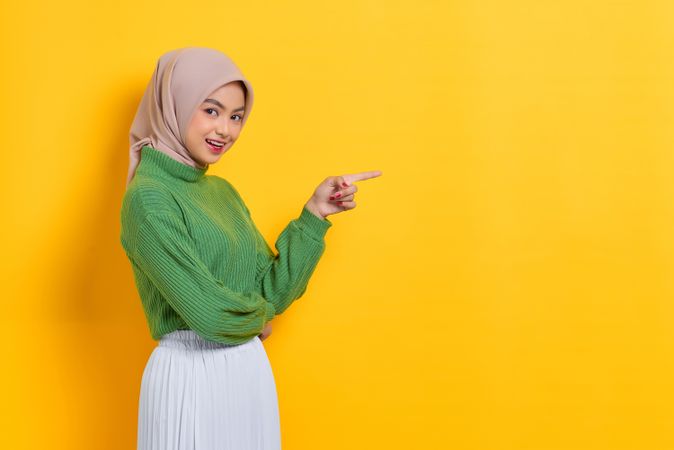 Woman in headscarf facing to the side and pointing at yellow copy space