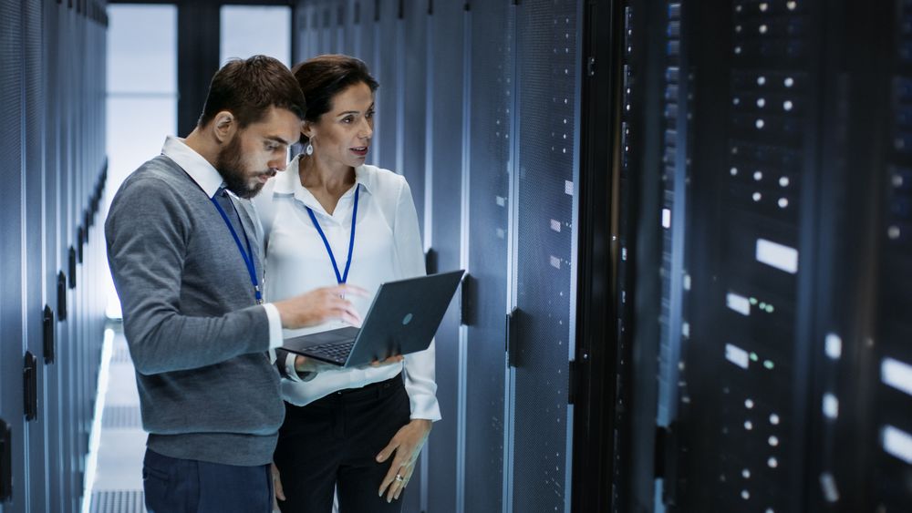 Male IT Specialist Holds Laptop and Discusses Work with Female Server Technician.