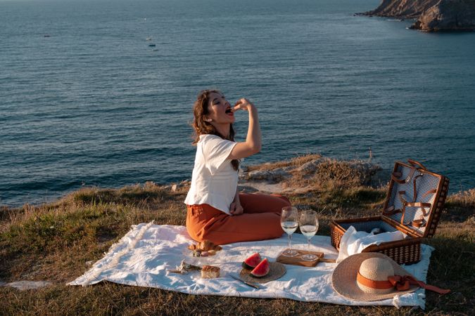 Woman with glass of drink on picnic mat near sea and mountains