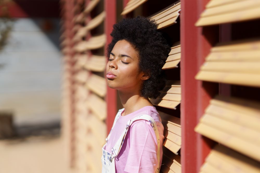 Female in overalls leaning on wooden shutters with face to the sun with eyes closed