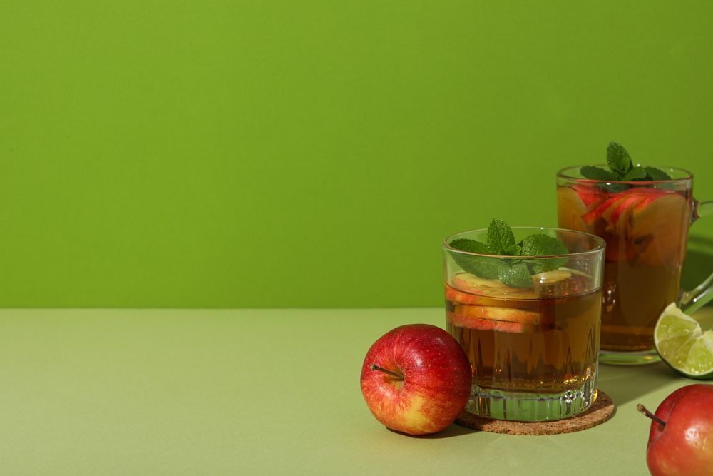 Glasses with apple cider, red apples and piece of lime on green background, space for text