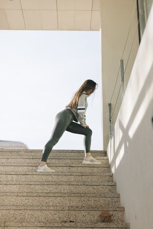 Full length side view shot of woman in grey workout pants stretching on stairs outside