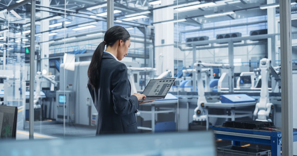 Southeast Asian Engineer Using Laptop Computer and Looking Around a Factory Facility with Equipment Producing Modern Electronic Components for Different Industries. Footage From the Back