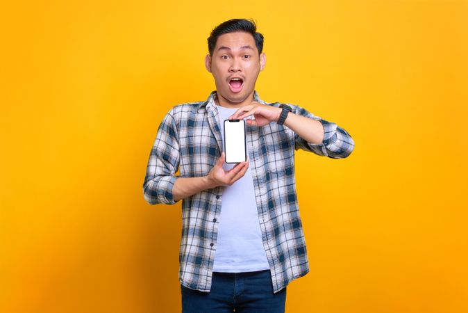Surprised Asian male showing blank screen of smart phone with both hands in studio shoot