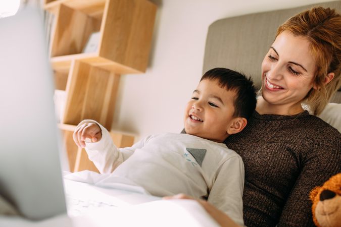 Smiling young woman with a little boy on the bed watching movies on laptop