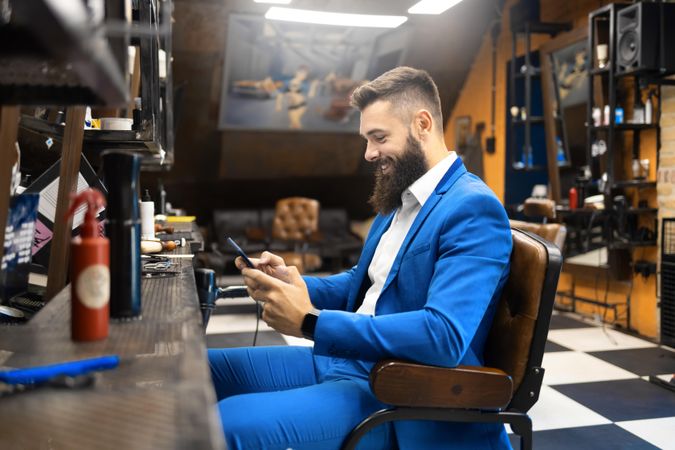 Smiling man in sharp blue suit checking phone in barber’s chair in salon