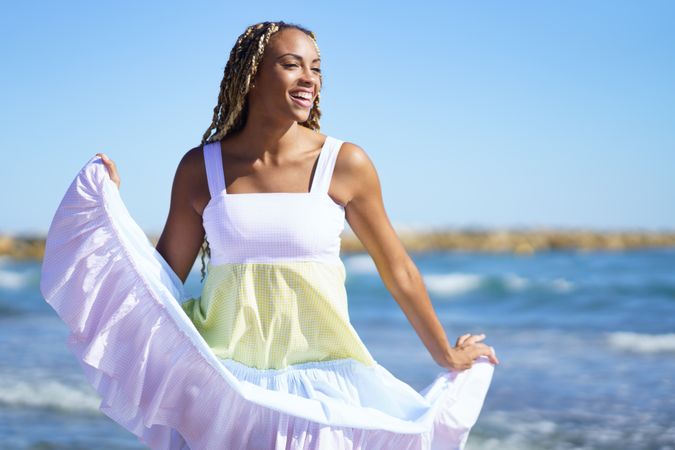 Black female holding up bottom of colorful dress while walking along the shore